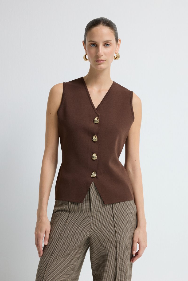 Donna che indossa un gilet senza maniche marrone con bottoni dorati decorativi e pantaloni a vita alta a quadri, in piedi davanti a uno sfondo bianco semplice.