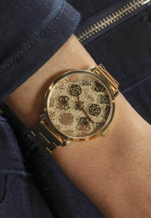 Orologio d'oro con un quadrante scintillante, accenti neri e motivi del logo; presenta un bracciale metallico e una cassa rotonda, visto su un polso.