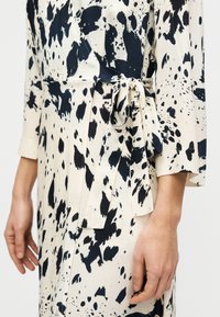 Κρεμ φόρεμα με μαύρο αφηρημένο σχέδιο κηλίδων, με σχέδιο τύπου wrap, μανίκια τριών τετάρτων και λεπτομέρεια στη μέση με δέσιμο. Ομαλή υφή υφάσματος.