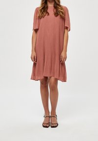 Růžové skládací šaty s krátkými rukávy, volným střihem a délce ke kolenům. Spárováno s černými sandály na ramínka.
