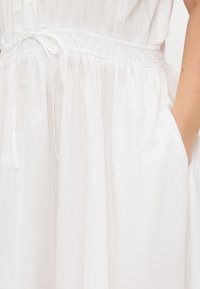 Weißes Baumwollkleid mit gerafftem Taillenbereich, Bindegurt und Seitentaschen. Sanfte Textur mit einem einfachen, fließenden Design.