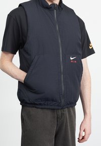 Mörkblå väst utan ärmar med texturerad tyg, dragkedja fram, sidofickor och en röd "AIR" logotyp; bärs över en svart t-shirt med kort ärm.