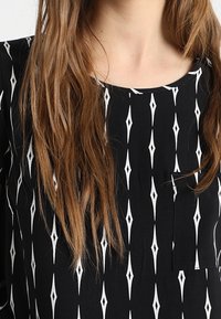 Svart topp med ett diamantmönster i vitt, rund halsringning och en liten ficka. Tyget verkar glatt och lätt.