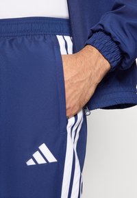 Survêtement de sport bleu marine avec des accents à trois bandes blanches ; comprend des poches zippées et des poignets élastiques. Le matériau semble lisse et léger.