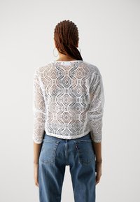 Hvit heklet topp med et blondeaktig mønster, trekvart ermer, kort design. Kombinert med høytlivs blå denimjeans.