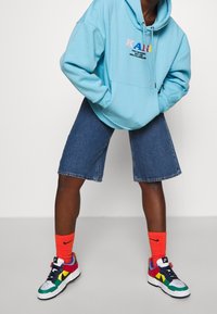 Sudadera azul con logotipo multicolor, pantalones cortos de mezclilla y zapatillas multicolores. Calcetines naranjas brillantes. Corte holgado, estilo casual.