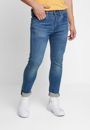 Jeans de mezclilla oscuros ajustados con un ligero desgastado, cinco bolsillos y puños enrollados, combinados con zapatillas blancas.