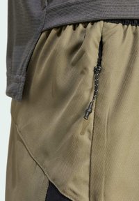 Pantalones cortos de exterior, tejido verde de textura suave, cinturilla elástica y detalles en negro. Incluyen un bolsillo lateral con cremallera y un tirador.