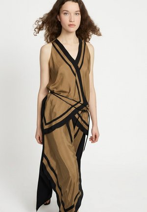 Frau mit lockigem Haar, die ein ärmelloses, braun-schwarzes asymmetrisches Kleid mit Wickeldetail und dünnen Taillenbändern trägt.