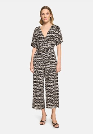 Femme portant une combinaison noire et beige à motifs géométriques, manches courtes, col en V, taille ceinturée et jambes larges raccourcies, associée à des sandales noires.