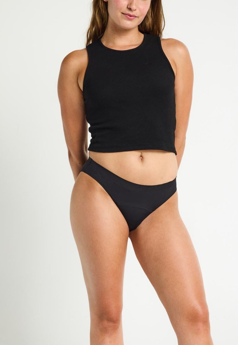 Une femme portant un crop top noir sans manches et des sous-vêtements taille haute noirs, se tenant avec les mains derrière le dos contre un fond blanc.