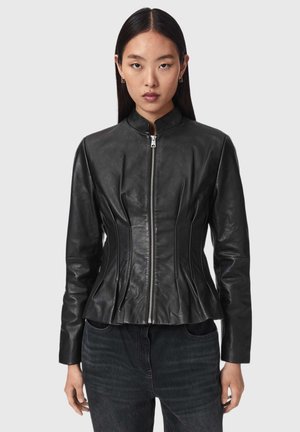Femme aux cheveux noirs et raides porte une veste en cuir noire ajustée avec une fermeture éclair à l'avant et un col montant, associée à un jean foncé.