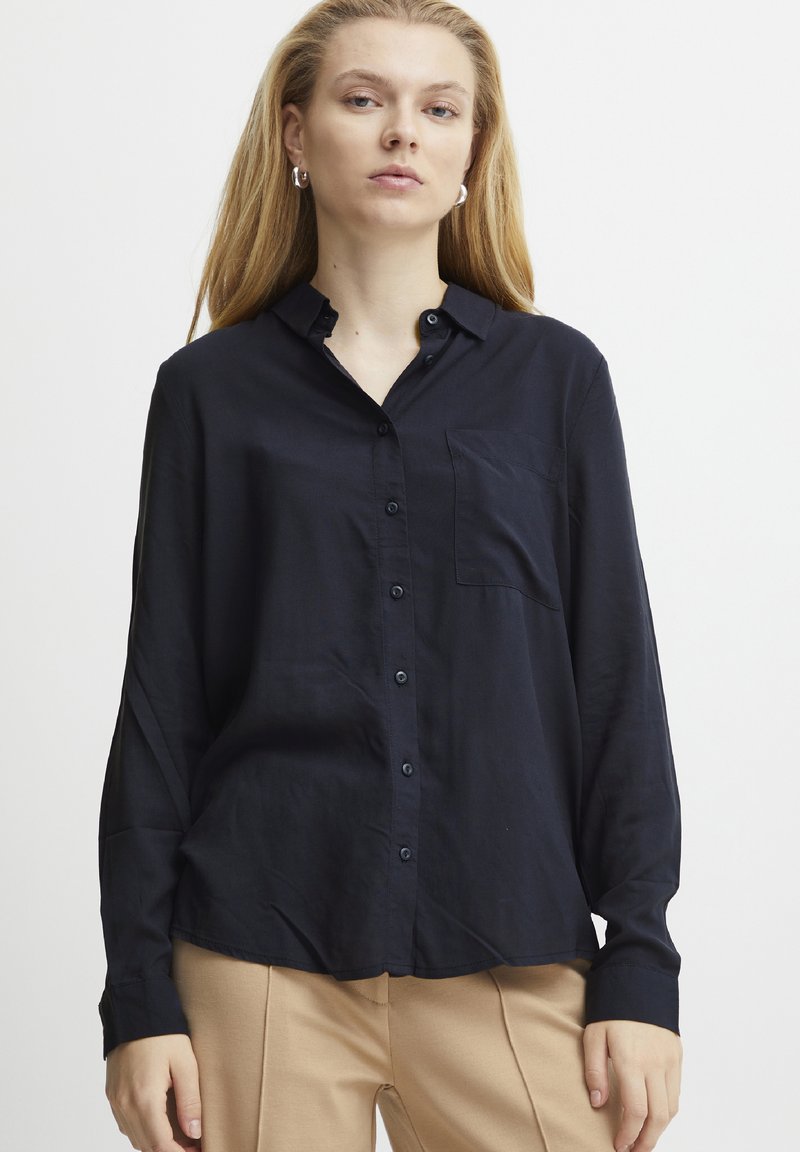 Camicia button-up blu navy con maniche lunghe, tasca sul petto e una texture morbida e liscia. Il tessuto sembra leggero e si drappeggia leggermente.