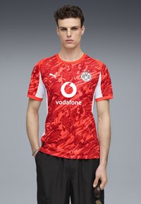 Rød og hvid kortærmet sportsjersey med abstrakte mønstre, der har et cirkulært Vodafone-logo og BVB-badge, lavet af letvægtsstof.