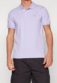 Polo en coton lilas avec col, patte de boutonnage à deux boutons et logo brodé "GAP" sur la poitrine. Manches courtes et coupe ajustée.