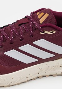 Chaussure de sport bordeaux avec un dessus en tissu texturé, des rayures blanches et une semelle beige marbrée. Présente des éléments en or sur la languette.