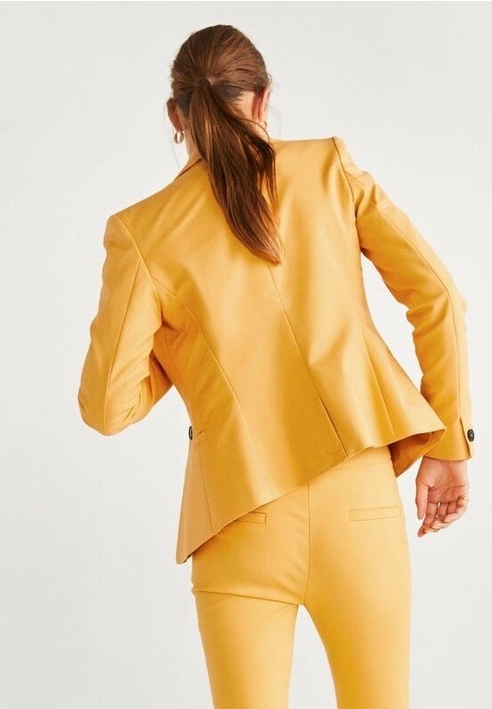 Femme aux cheveux bruns attachés en queue de cheval, portant un blazer cintré jaune moutarde et un pantalon assorti, vue de dos sur un fond uni.