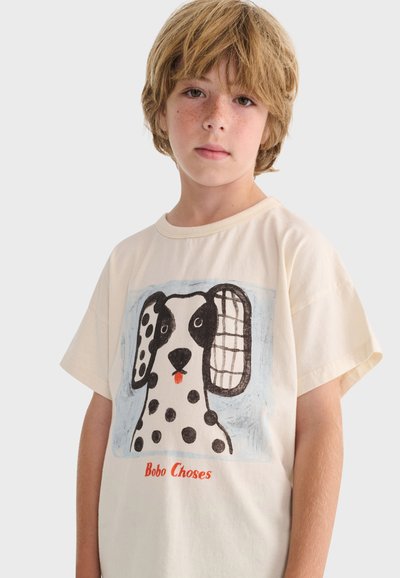 Ragazzo giovane con capelli castano chiaro che indossa una maglietta bianca con l'illustrazione di un cane macchiato e la scritta "Bobo Choses" su uno sfondo semplice.