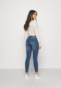 Fitted, high-waisted blue jeans with back pockets, paired with a cream long-sleeve sweater and light-colored sneakers on a white background.