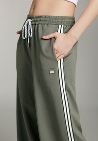 Grüne Sporthose mit Zugbandbund, Seitentaschen, weißen Streifen an den Seiten und einem kleinen Logo-Patch am Bein.