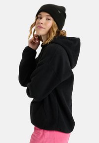 Felpa con cappuccio nera in pile, vestibilità oversize e tasche frontali. Indossata con un berretto di lana nero. Il modello posa di lato su uno sfondo bianco.