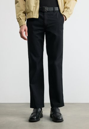 Άνδρας που φοράει μαύρο παντελόνι κανονικής γραμμής, μαύρη δερμάτινη ζώνη, μαύρα μοκασίνια, μπεζ σακάκι, με τα χέρια στις τσέπες, μπροστά σε απλό φόντο.