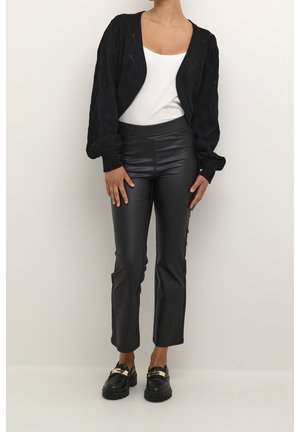 Vrouw draagt een wit topje, een zwarte gebreide open cardigan, zwarte nepleren broek en zwarte loafers met gouden accenten.