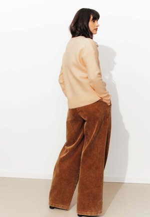 Pull léger pêche avec bord côtes, associé à un pantalon en velours côtelé brun à jambes larges. Texture lisse, design minimal. Le modèle pose avec les mains dans les poches.