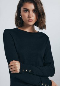 Maglione nero-knit con scollo rotondo, polsini a coste con tre bottoni dorati e una superficie liscia. Design aderente.