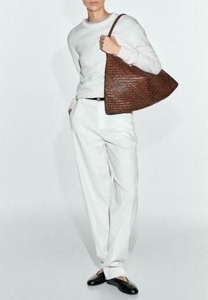 Osoba v bílém svetru a kalhotách, černém pásku a botách, nesoucí velkou hnědou pletenou ramenní tašku.