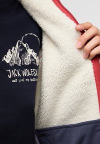 Marineblaue Fleecejacke mit einem weißen, weichen Innenfutter, das ein Bergmotiv und Text zeigt. Rote Akzente entlang des Reißverschlusses und der Kanten.