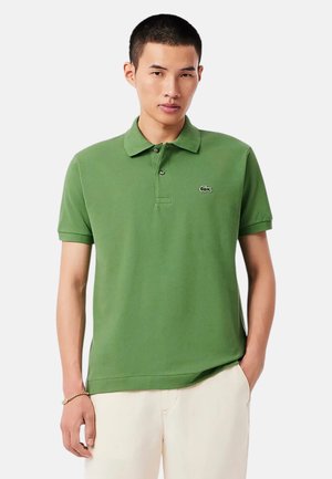 Hombre con camisa polo de manga corta verde con pequeño logo de cocodrilo, pantalones beige y pulsera dorada, de pie con una mano en el bolsillo.