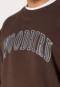 Sudadera marrón con texto blanco "WOODBIRD" en diseño arqueado. Cuello acanalado y textura de tela suave. Diseñada con un ajuste relajado.