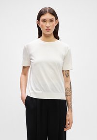 Top blanco de manga corta en tela suave, con cuello redondo y dobladillo acanalado. El modelo lo lleva con pantalones negros, mostrando un ajuste casual.