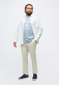 Άνδρας με πλεξούδες, ντυμένος με ανοιχτή λευκή πουκάμισα πάνω από γαλάζιο μπλουζάκι με στάμπα, μπεζ παντελόνι και μαύρα αθλητικά παπούτσια, στέκεται μπροστά σε ένα μονόχρωμο φόντο.