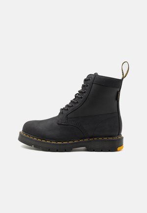 Bottine noire à lacets avec semelle épaisse, surpiqûres jaunes et boucle en tissu à l’arrière, sur un fond clair uni.
