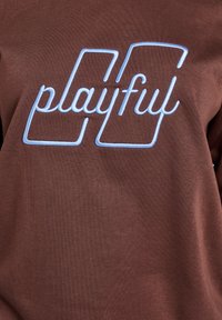 Rudos spalvos džemperis su šviesiai mėlynu siuvinėtu paveikslėliu „playful“. Minkštos tekstūros, laisvo silueto ir minimalistinio dizaino.