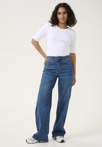 Jean en denim bleu taille haute avec un design à jambes larges, associé à un haut ajusté à manches courtes en rib de couleur blanche. Accentué par un bouton en argent.