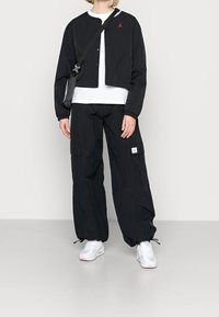 Svart kort jacka med röd logga, vit t-shirt under, och svarta cargobyxor. Vita sneakers med rosa detaljer.