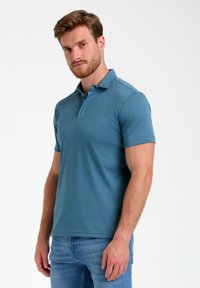 Kurzarm-Poloshirt in gedämpftem Blau, aus glattem Stoff, mit einem knöpfbaren Kragen und subtiler Logo auf der Brust. Getragen mit hellen Jeans.