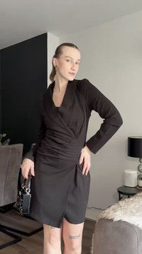 Svart omslagkjole med lange ermer, teksturert stoff og samlet detaljering i midjen; kombinert med en liten svart håndveske.