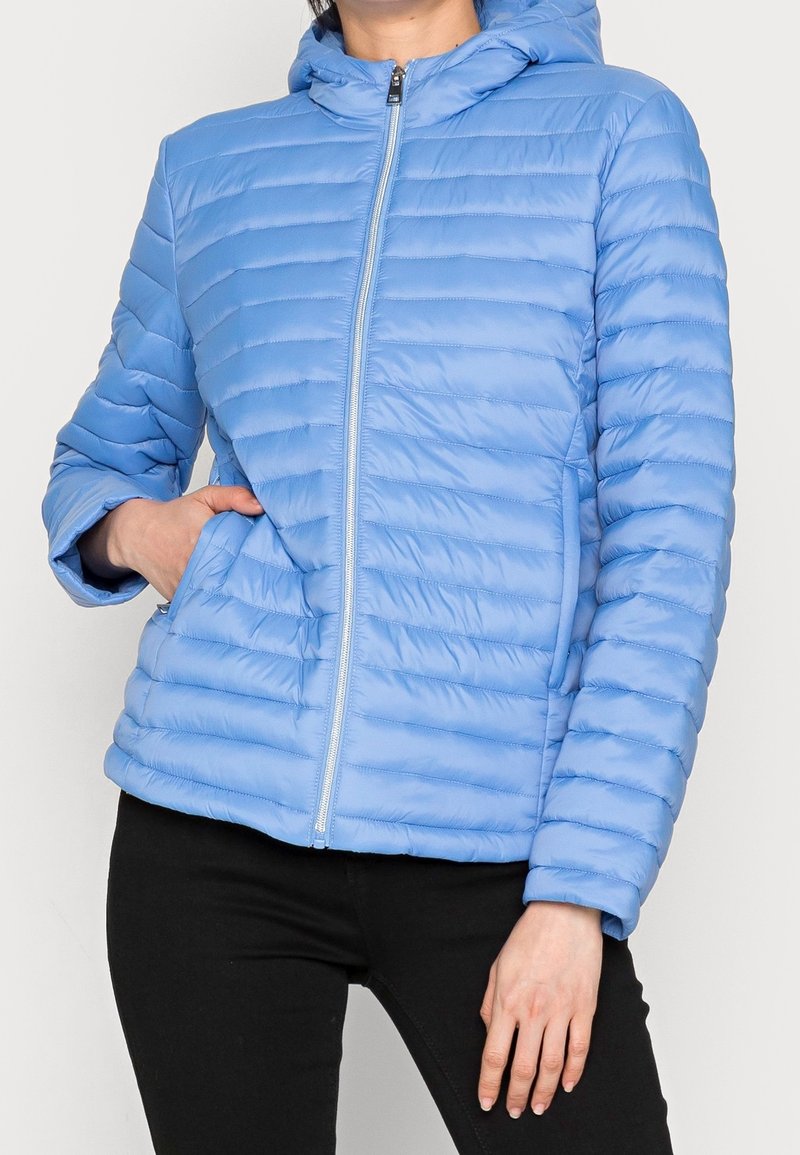 Veste matelassée à capuche de couleur bleu clair avec des coutures verticales, fermeture éclair et poches latérales. Fabriquée en matériau lisse et rembourré.