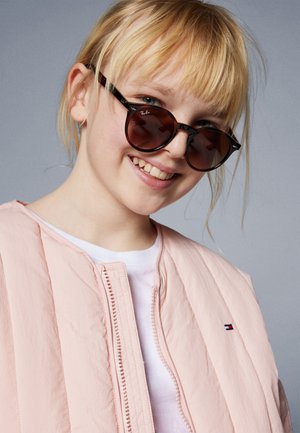 Gafas de sol en forma de ojo de gato de color negro y carey con lentes oscuros, combinadas con una chaqueta bomber de color rosa claro con cierre de cremallera y detalle de logo.