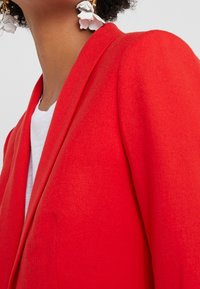 Punainen bleiseri, jossa on teksturoitu kangas, huippukorot ja rakenteelliset harteet. Päällä valkoinen paita, tyylitelty koristelu korvarenkailla.