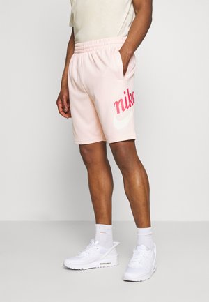 Mies, joka pukeutunut vaaleanpunaiseen Nike-shortsiin, jossa on punainen logo, ja valkoisiin lenkkareihin, seisoo yhdellä kädellä taskussa yksinkertaisen taustan edessä.