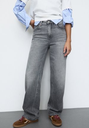 Persoon draagt vervaagde zwarte wijdere grijze jeans, bruine sneakers met rode veters, een wit topje over een blauw gestreept shirt, staat met één hand in de zak.