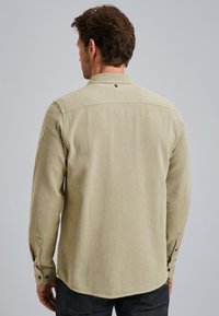 Chemise khaki à manches longues, en tissu texturé, avec un empiècement aux épaules et des poignets boutonnés. Présente une patch sur la manche gauche.