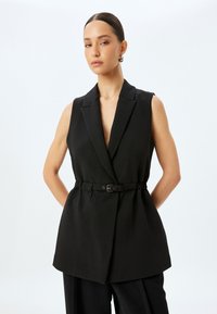 Blazer senza maniche nero con scollatura a V profonda, vestibilità sartoriale e dettaglio con cintura in tessuto. Texture liscia, forma strutturata e design minimalista.