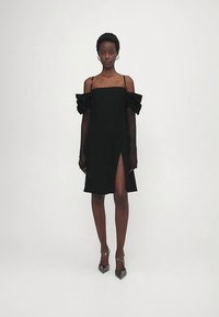 Woman wearing a black off-shoulder dress with ruffled sleeves, mesh long gloves, and black pointed heels, standing against a plain background.