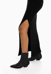 Zwarte geribbelde jurk met een hoge split tot boven de dijen, gecombineerd met zwarte enkellaarsjes met een spitse neus en een lage blokhak.